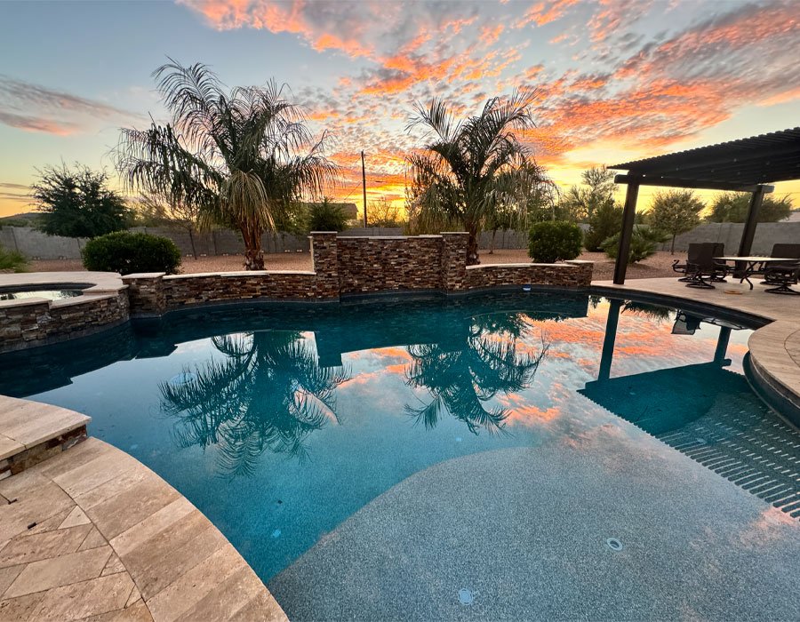 Luxurious backyard pool with clear water reflecting a vibrant sunset, surrounded by palm trees and a stone wall, showcasing a serene outdoor space ideal for relaxation and pool maintenance services.