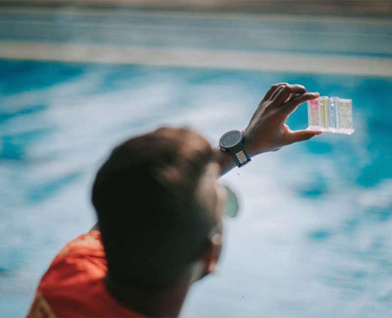 Person testing pool water with chemical test strips, pool water in background, emphasizing visual pool inspections in Tucson.