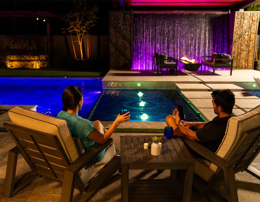 Couple relaxing by illuminated pool with modern lighting features, showcasing vibrant colors and an inviting atmosphere for nighttime enjoyment.