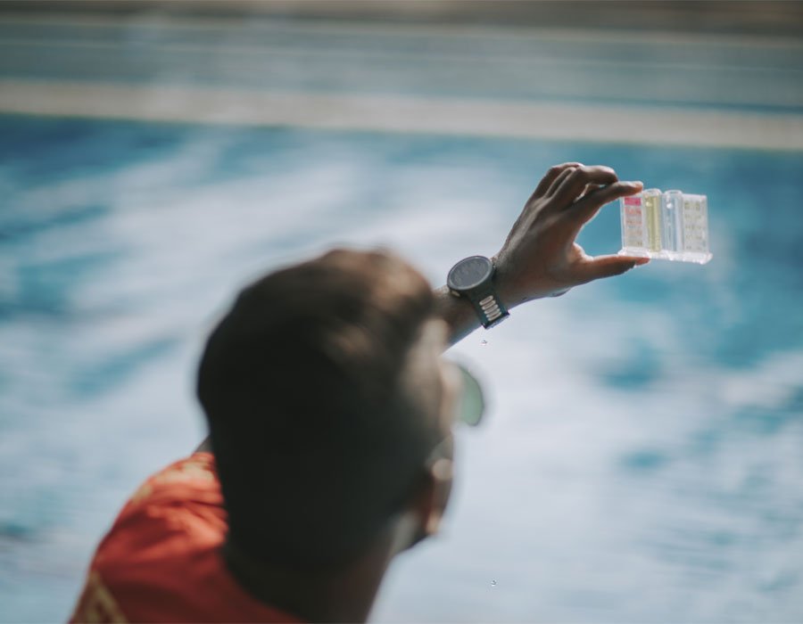 Person testing pool water quality with a water testing kit, showcasing color indicators for chemical balance, with a pool in the background.