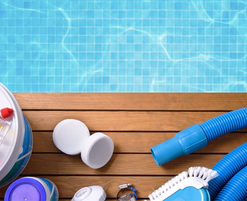 Pool maintenance tools including chemical containers, a vacuum hose, and cleaning brushes on a wooden deck beside a clear blue swimming pool, emphasizing professional weekly pool service.