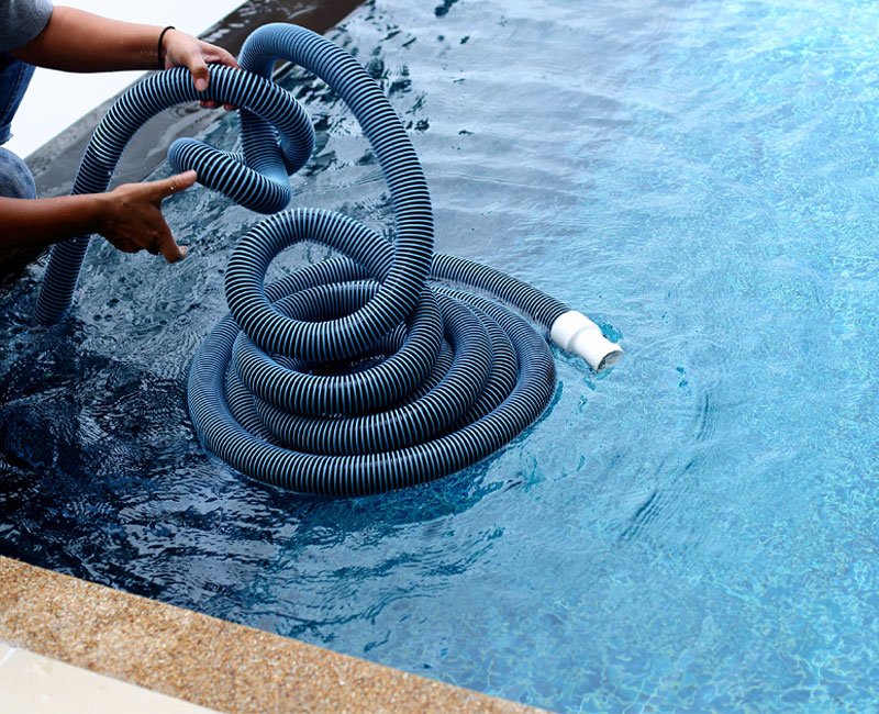 Pool maintenance technician handling a blue pool vacuum hose in clear water, emphasizing professional weekly pool service for clean and safe swimming environments.