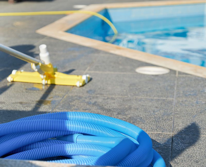 Pool maintenance equipment including a yellow vacuum head and blue hose, positioned near a swimming pool, illustrating professional cleaning service for optimal water chemistry and cleanliness.